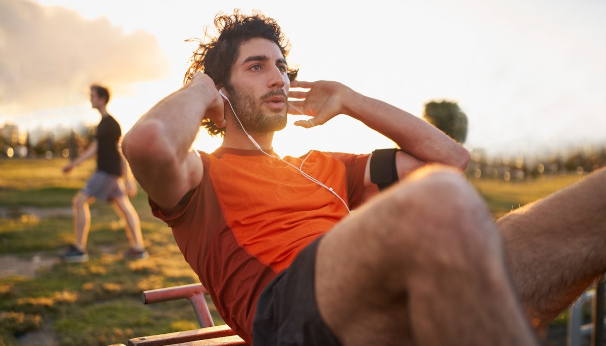 Confident young man listening to music in headphones from smart phone armband doing exercises on public equipment in the outdoor gym at the park