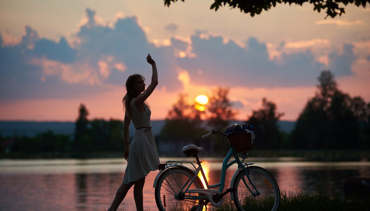 Tender woman in a dress posing with a vintage bicycle near the river against the backdrop of a bewitching sunset. Warm evening in the nature. Freedom lifestyle